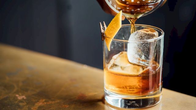 A glass tumbler with ice and amber whiskey is being poured with a garnish of orange peel, a lemon twist, and a fancy swirl.