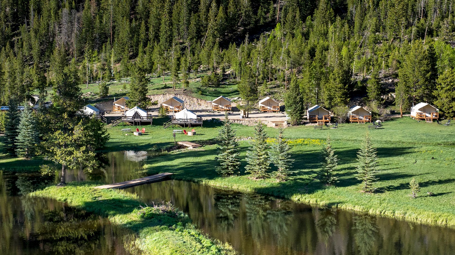 A tranquil lakeside village with wooden cabins among pine trees, lush green grass, and a calm reflective water body.