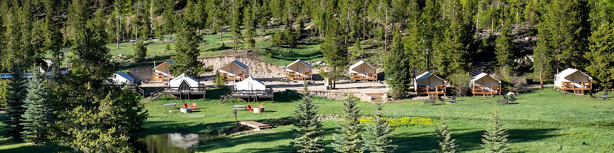 A tranquil lakeside village with wooden cabins among pine trees, lush green grass, and a calm reflective water body.