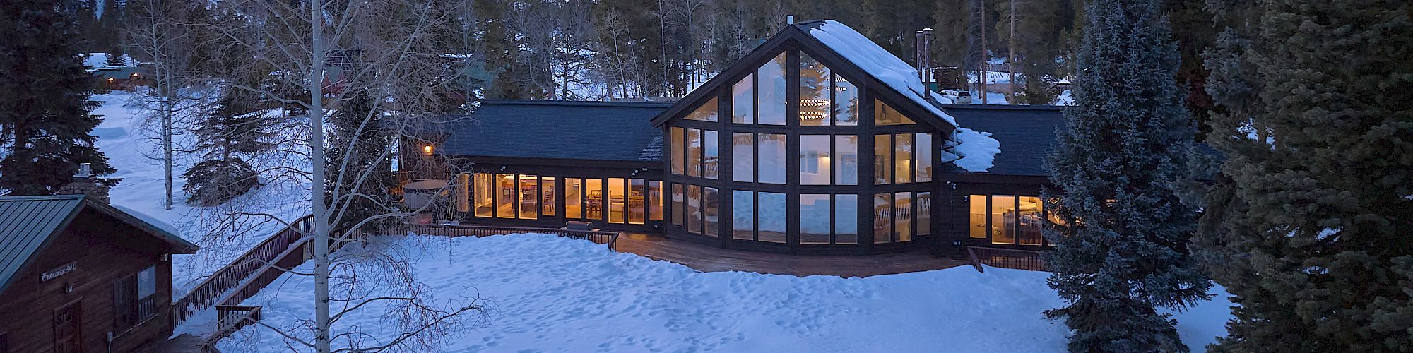 A modern lodge sits in a snowy mountain setting, warm lights glowing from large windows as twilight settles over snow-covered trees and hills.