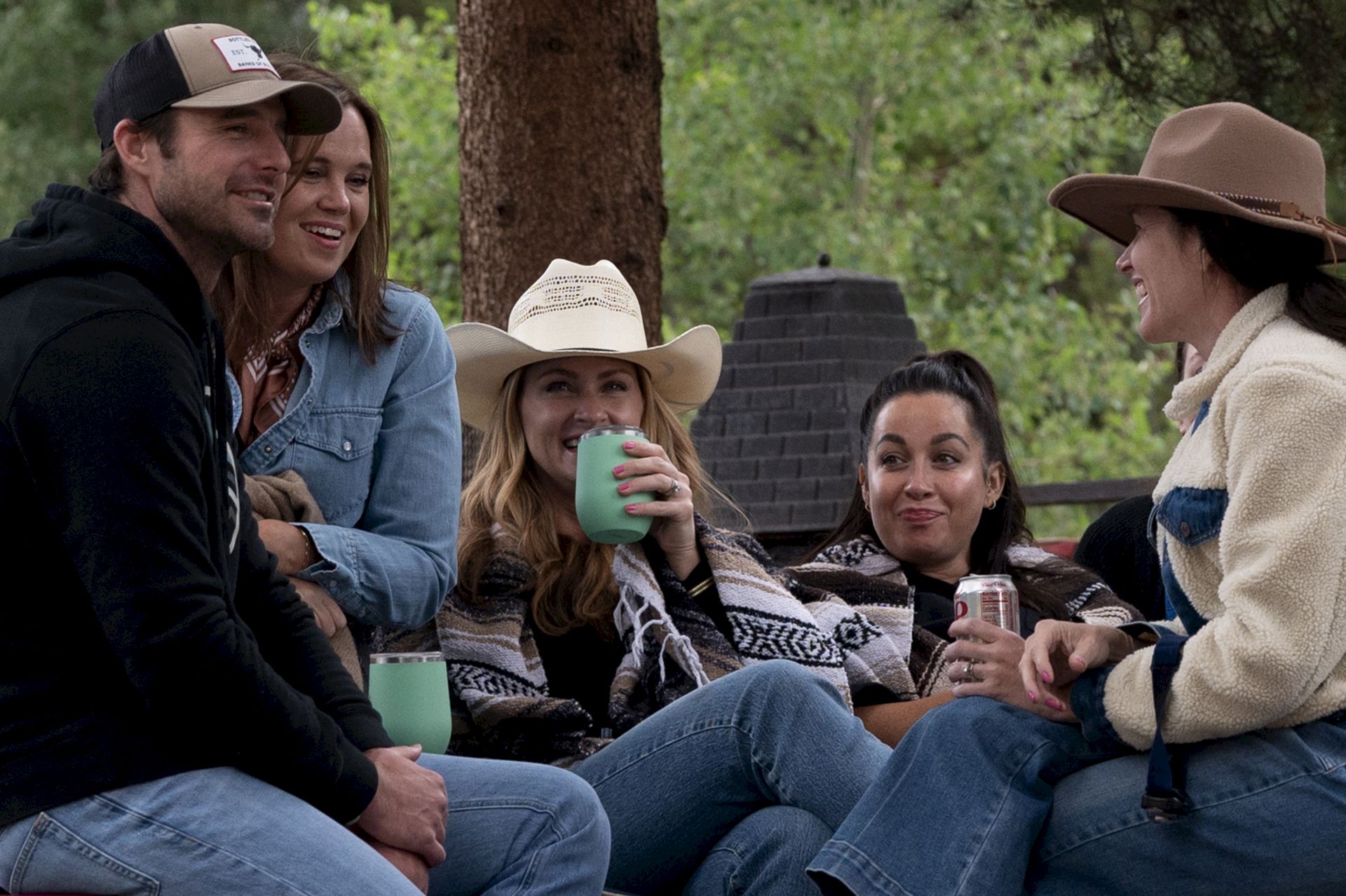 A group of six friends sits outdoors around a campfire area, smiling and sipping drinks, wearing casual outdoor outfits and hats.