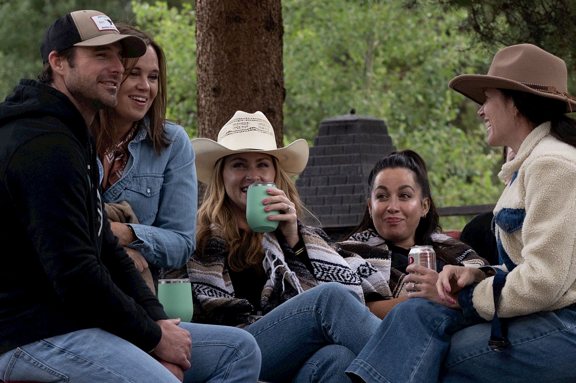 A group of six friends sits outdoors around a campfire area, smiling and sipping drinks, wearing casual outdoor outfits and hats.
