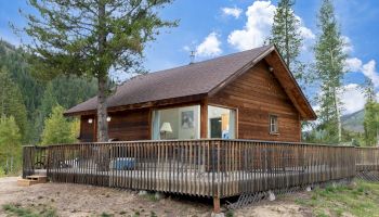 A log cabin with a wide wooden deck sits on a hillside, surrounded by trees and mountains under a blue, partly cloudy sky.