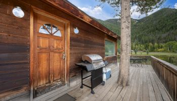 A wooden cabin with a front door, grill on a stand, a tree by a wooden deck, and a scenic mountain lake backdrop.