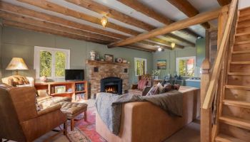 Cozy rustic living room with a stone fireplace, wooden beams, and comfy seating; stairs on the right, bright windows, and a warm, inviting vibe.