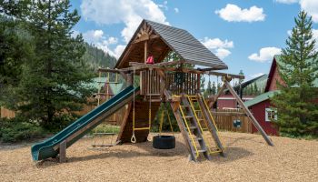 A wooden playground with slides, climbing ladders, a tire swing, and a shaded fort, set among trees under a blue, partly cloudy sky.