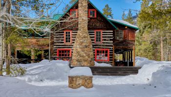 A log cabin with a tall stone chimney in a snowy yard, evergreen trees surrounding, and a stone bench in front.