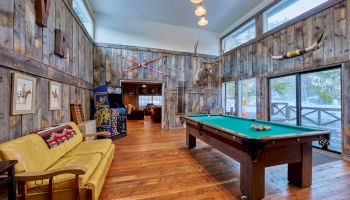 A rustic living room with wood plank walls, yellow sofa, pool table, framed art, mounted weapons and decor, and large windows overlooking a deck.