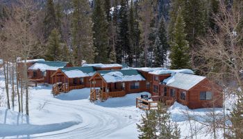 Snowy log cabins nestled among pine trees in a mountain setting, with fresh tracks in the snow and a serene winter scene.