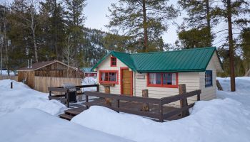 A small cabin with a green metal roof sits among snowy pines, fenced porch, and a wooden picnic table, in a wintery woodland setting.