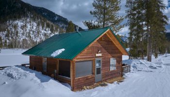 A small wooden cabin with a green metal roof sits in a snowy landscape, surrounded by tall trees and distant mountains.