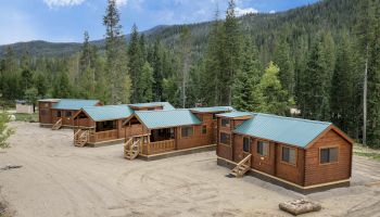 A row of wooden cabins with green roofs sits in a forested mountain clearing, with dirt ground and trees in the background.