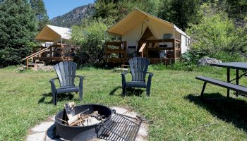 Two campground tents set in a grassy clearing with wooden decks, two blue chairs, a fire pit, and a picnic table, surrounded by pine trees.
