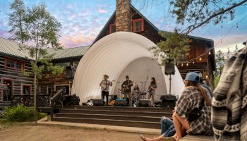A small band plays on a curved white stage at an outdoor venue with a rustic wooden building, while a seated audience enjoys the sunset.