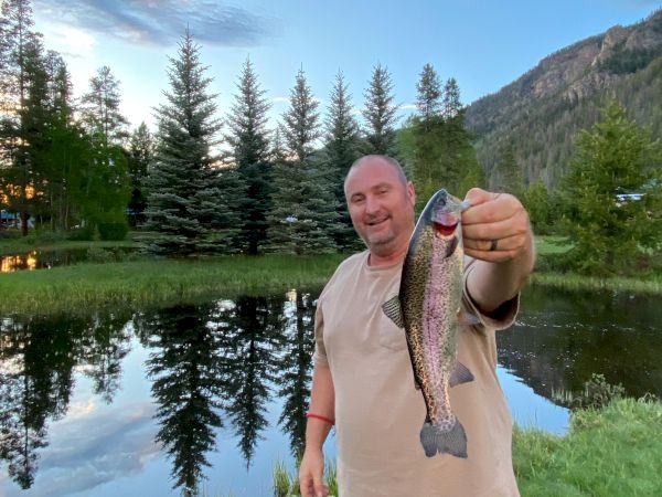 A man stands by a tranquil lake in a scenic mountain setting, proudly holding a small rainbow trout toward the camera. The sky is clear, reflections mirror the trees, and he wears a beige shirt and dark pants.