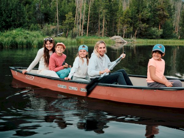 Five people ride a long red boat on calm water, smiling near a forested shoreline as they paddle together, wearing hats and casual clothes.