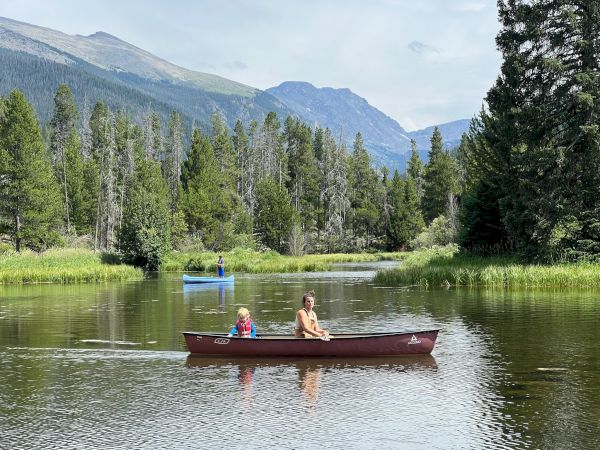 Two people row a small red canoe on a calm lake surrounded by pine trees and towering mountains in the distance.