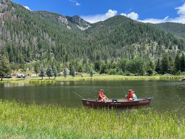 Two people fishing from a red boat on a calm lake, surrounded by grassy banks and forested mountains under a blue sky.