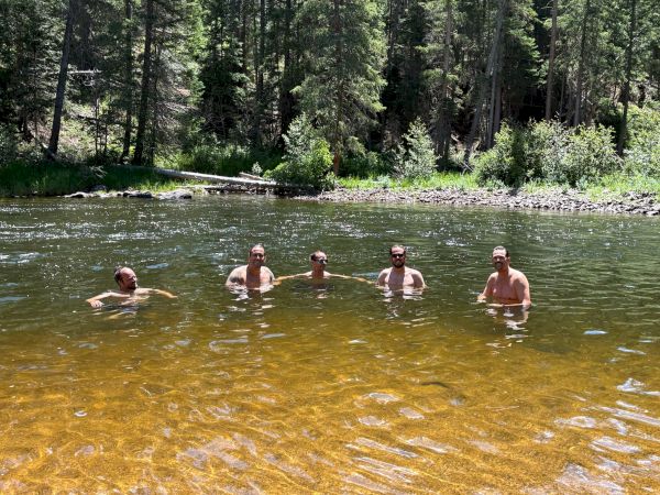 Five people stand chest-deep in a clear river, forest backdrop, sunlit water, casual summer outing, relaxed group swim.