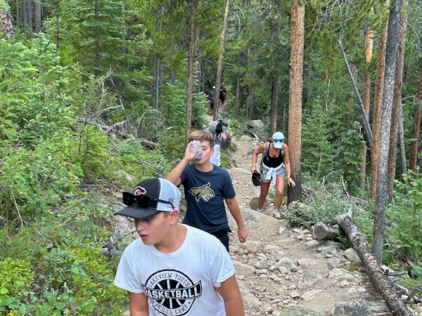 A group of hikers traverses a rocky forest trail, one wearing a white shirt at the front, surrounded by tall trees and greenery.