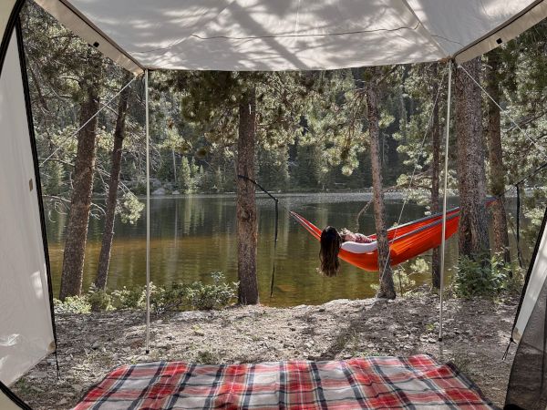 A view from a tent overlooking a lake with a hammock in trees and a red plaid blanket inside.