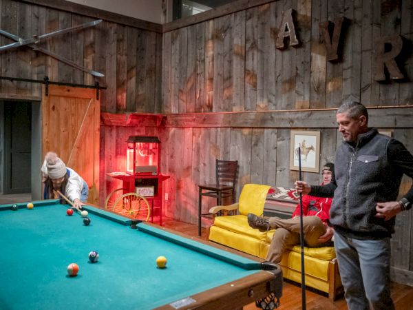 A cozy rustic game room with a pool table, red ambient light, a man lining up a shot, another man on the couch, and wooden walls.