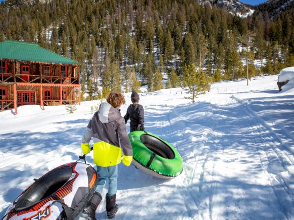 Two people in a snowy alpine setting with snow tubes (one black/red, one green) near a wooden lodge; pine trees and a clear sky in the background.