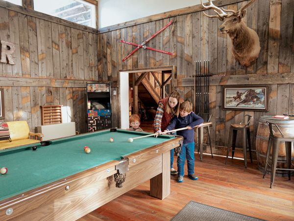 Two men play pool in a rustic wood-paneled room with deer antler decor, mounted deer head, and vintage bar stools nearby.