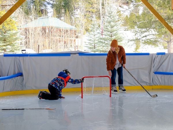 Two people on an ice rink playing street hockey; one kneels with a stick, the other stands near a small net under a wooden shelter.