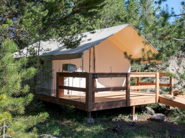 A canvas tent cabin on a wooden platform in a forest, with a railing and small deck, surrounded by trees and rocky ground.