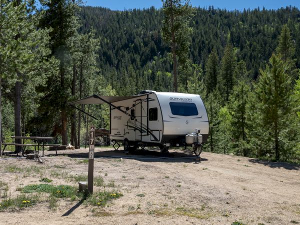 A white RV trailer parked on a dirt clearing under tall pines, with a small awning, picnic table, and forested hills in the background.