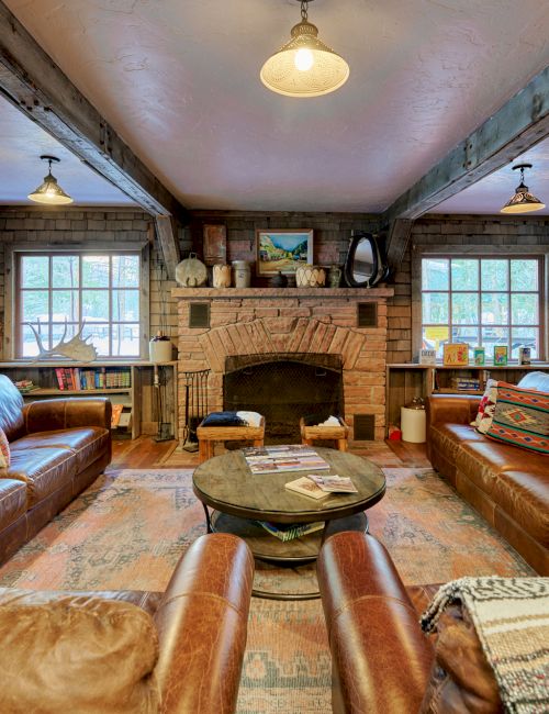 Cozy rustic living room with leather sofas, a stone fireplace, wooden beams, warm lighting, and a round coffee table in front of a brick hearth.