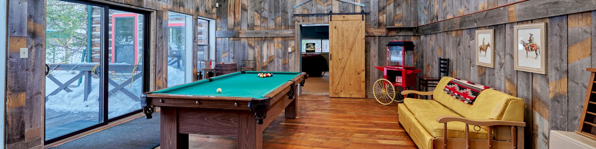 Cozy rustic game room with a pool table, wooden plank walls, a yellow couch, vintage decor, and large windows letting in natural light.