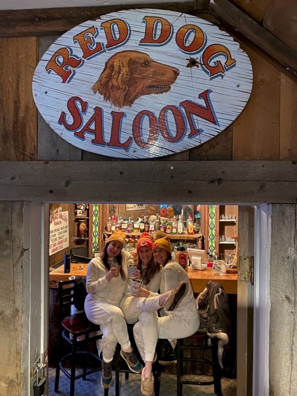 Three people in white outfits sit on stools under a rustic &ldquo;Red Dog Saloon&rdquo; sign, inside a cozy, vintage bar setting.