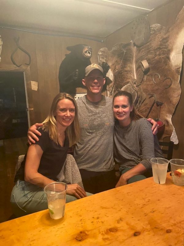 Three friends posing at a wooden bar, smiling with arms around each other in a cozy, rustic setting.
