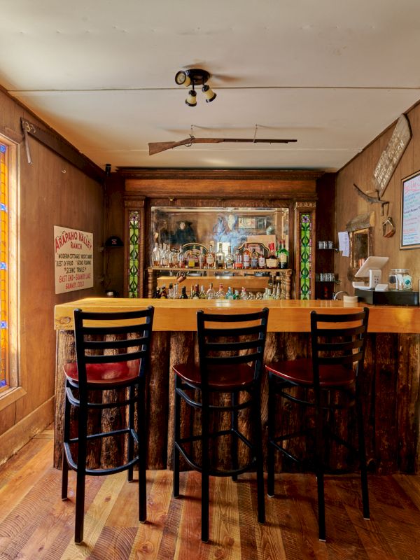 A cozy rustic pub interior with a wooden bar, high stools, warm lighting, stained walls, and a back bar stocked with bottles.