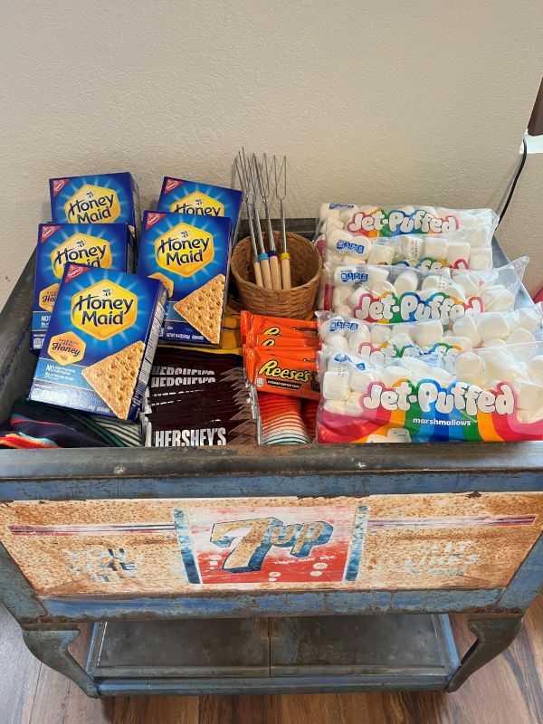 A small cart with snacks: multiple Honey Maid graham packs, Hershey&rsquo;s bars, a row of bagged candies (possibly Sour Patch/Delites), and a container with chopsticks on a tray.
