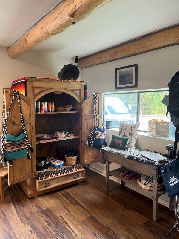 Cozy rustic room with wooden beams, a tall armoire filled with trinkets, a bench by a window, and hiking gear hanging on the wall.