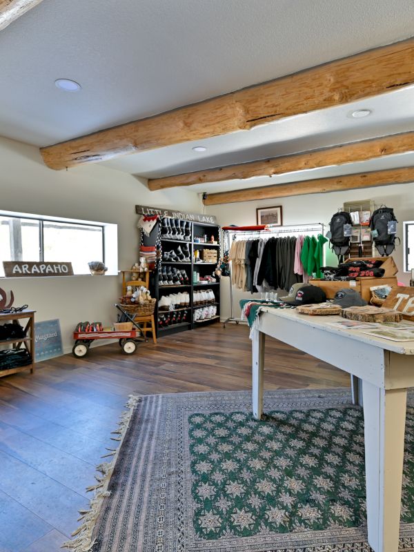 A cozy, bright thrift shop or studio with wooden beams, a white table centerpiece, clothing racks, colorful textiles, and vintage decor under skylight windows.