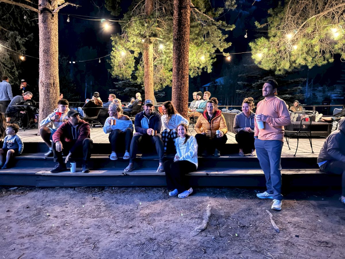 A group of people outdoors at night, sitting on a wooden platform under string lights among trees, chatting and relaxing by a campfire setting.
