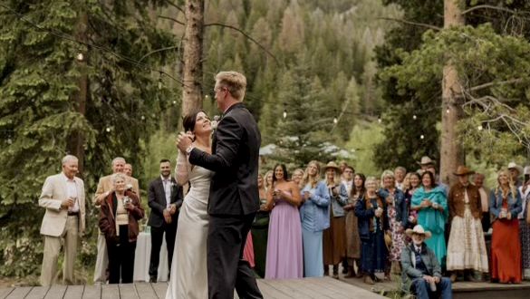 A couple dances a wedding first dance on a wooden deck, surrounded by guests in formal attire under tall trees in a forested setting.
