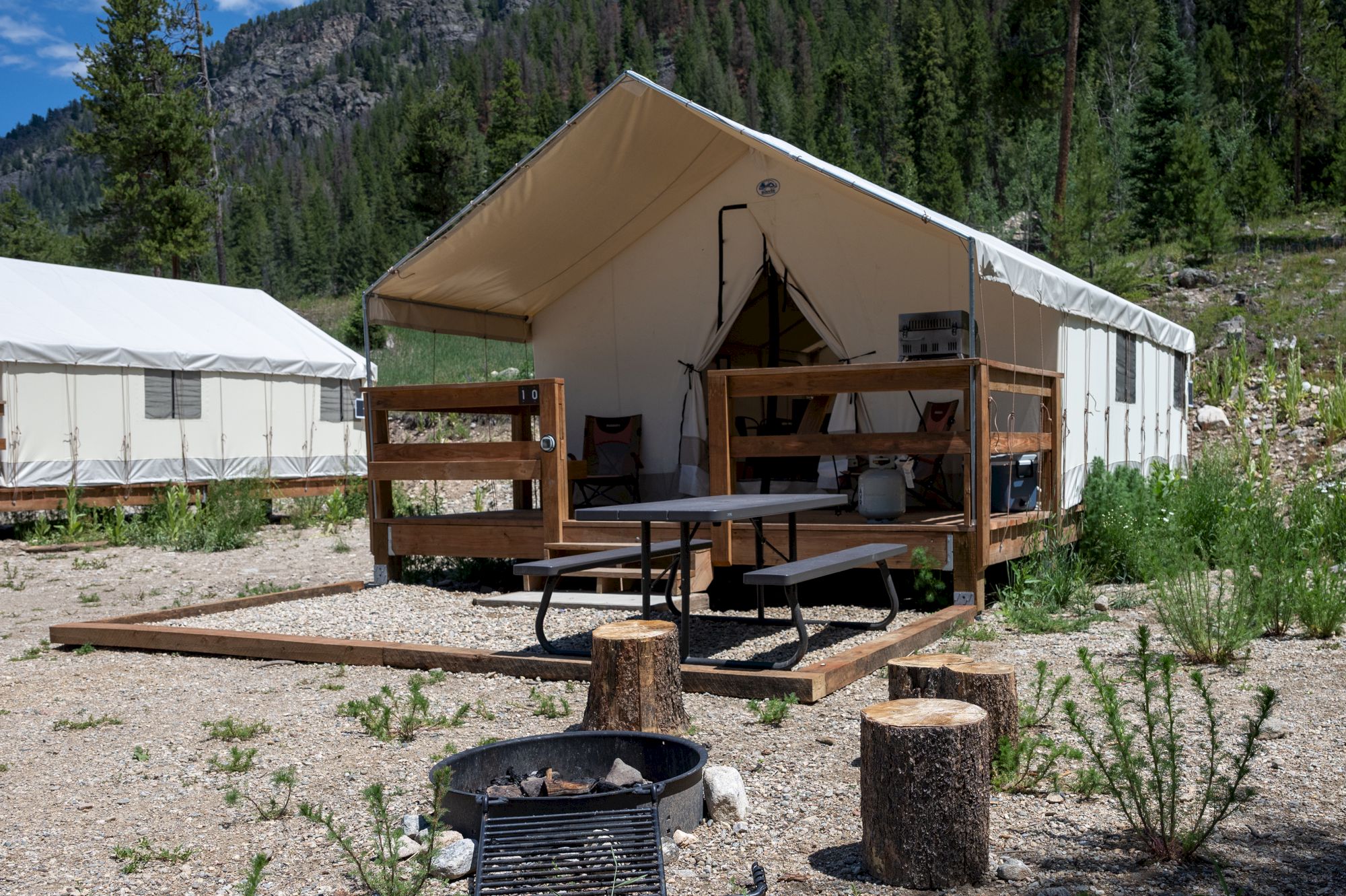 A campsite with a beige canvas tent, wooden deck, picnic table, and chairs, set in a rocky, forested area with stumps and a fire pit.