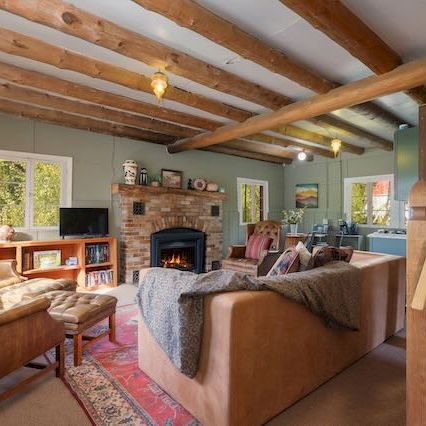 Cozy rustic living room with stone fireplace, wooden beams, comfy sofas, armchairs, coffee table, rug, lamps, stairs to the right, airy windows.