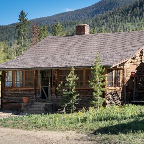 A rustic log cabin in a forested mountain setting, with a stone chimney, wooden porch, and surrounding trees and greenery.