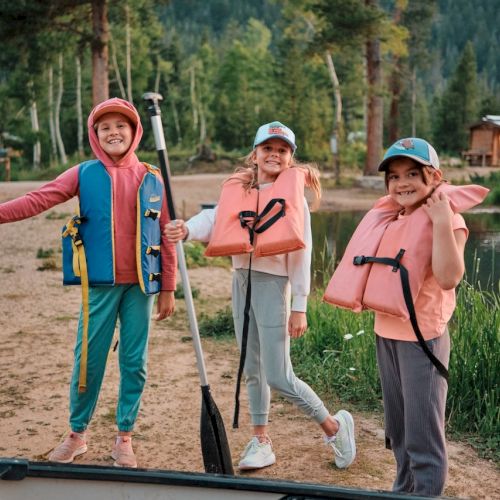 Three kids wearing life vests stand by a lake with paddles, ready for a boating outing at a lakeside campsite.
