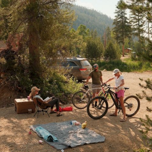 People relaxing outdoors by a rocky campsite: two on bikes with a car and trees in a mountain setting, a tarp on the ground and a seated couple.