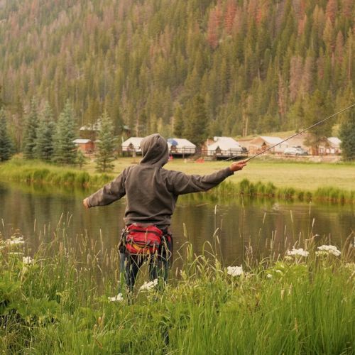 Two people fishing near a lake with tall grasses, trees, and houses in the background; mountains and a calm, rural scene.