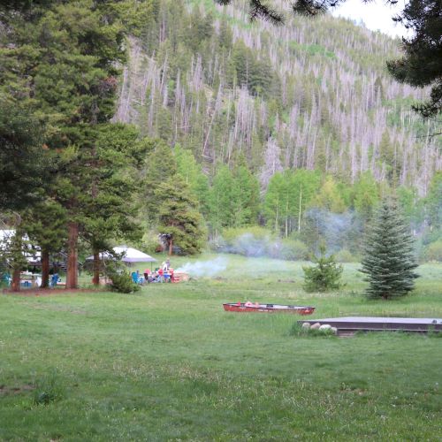 A grassy campsite under trees with a small group by a fire, smoke rising near tents, mountains in the background.