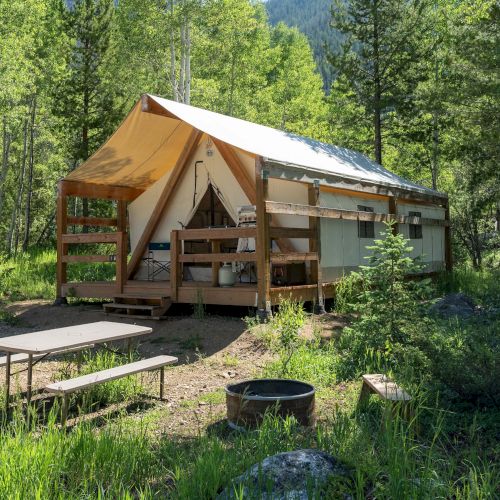 A canvas-roofed tent cabin in a forest clearing with a picnic bench, a fire pit, and surrounding green trees and sunshine.