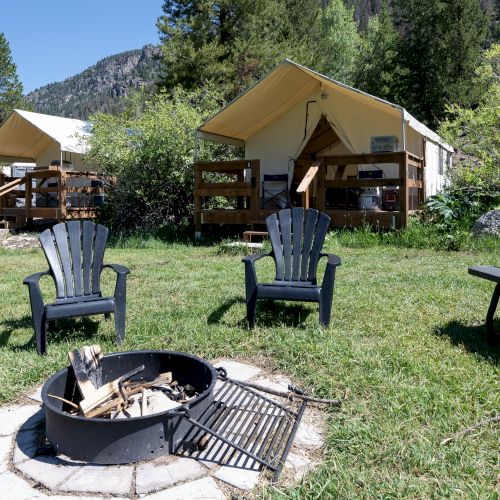A campsite with two tents, wooden porches, two blue chairs, a fire pit, and a picnic table on a grassy clearing in a forested area.